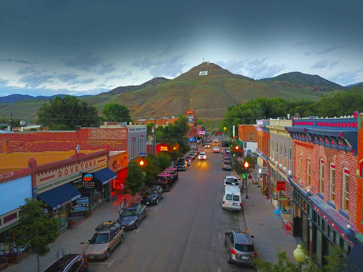 Downtown Salida at dusks Western buildings lights have started to come on with cars in the street. In the distance green covered hills meet a light-blue sky.