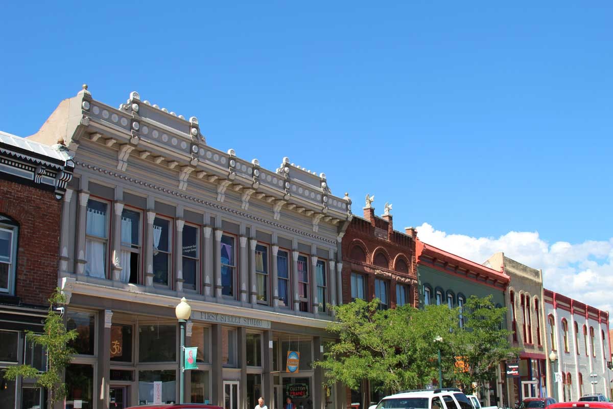 Two-story Victorian buildings sit on a street with a green tree below a blue sky.