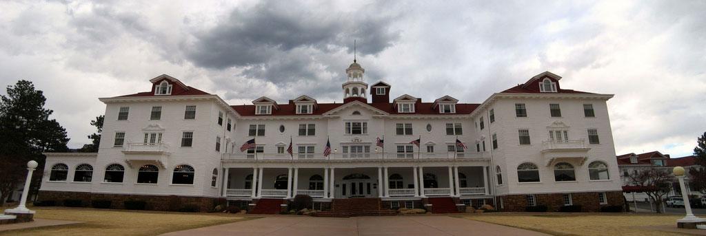 A white colonial-style building with a red roof and wide, pillar-framed front porch stands tall against a cloudy gray sky in Estes Park, Colorado.