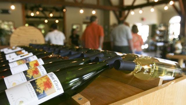 Rows of wine in green and clear bottles lay on a wooden rack at a winery in Colorado. People mill about in the background and strings of light twinkle above them.