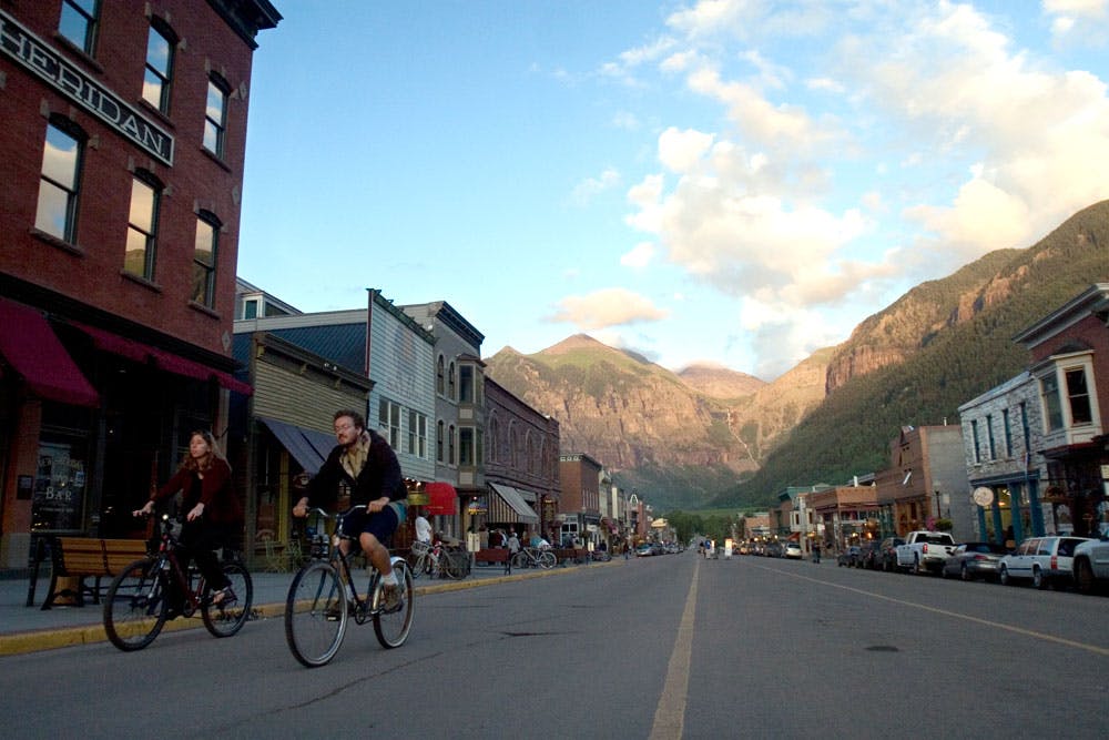 A couple rides on bikes down a peaceful street in Telluride, Colorado. Behind them historic buildings line the street and towering peak rise up near town.