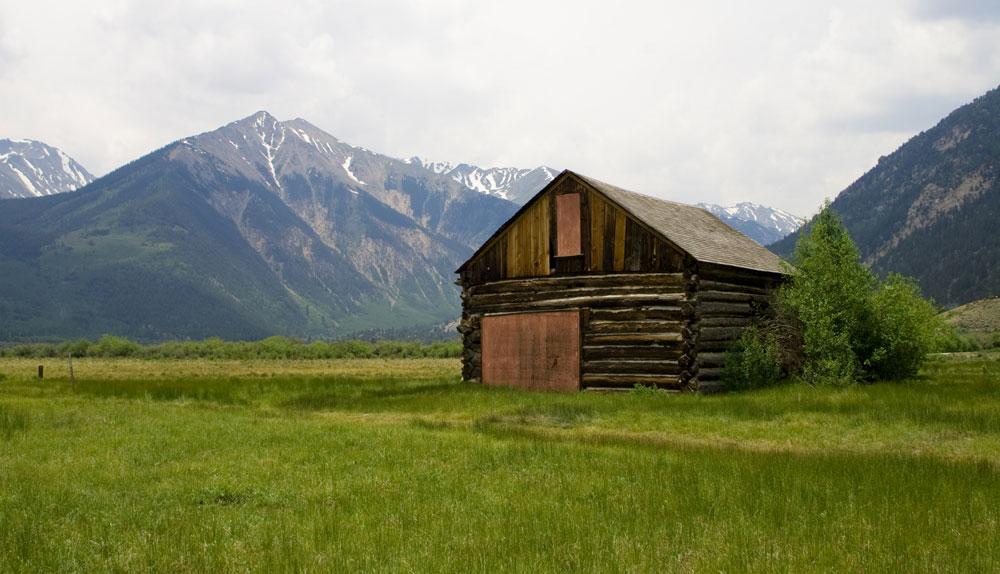 A boarded up wood cabin sits in a green meadow on a foggy day