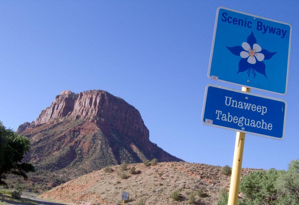 A red-rock butte pictured against a blue sky; a sign reads "Scenic Byway, Unaweep Tabeguache"