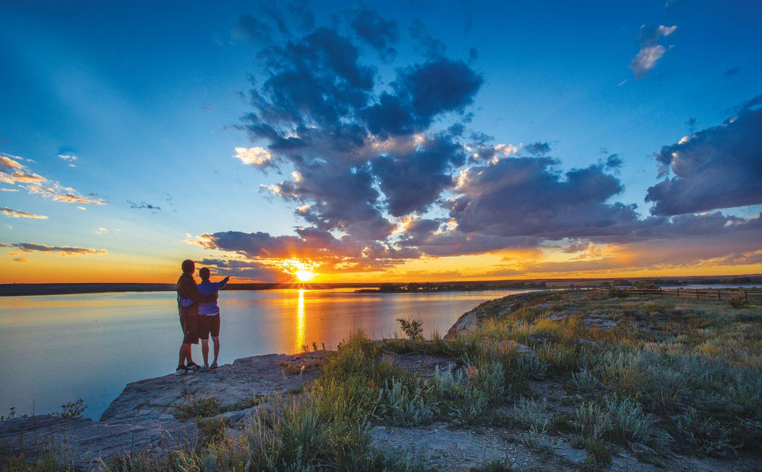 A couple embrace with one member pointing at the sunset across a lake.