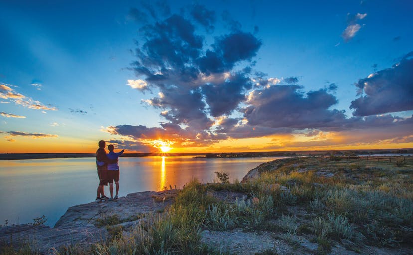 A couple embrace with one member pointing at the sunset across a lake.