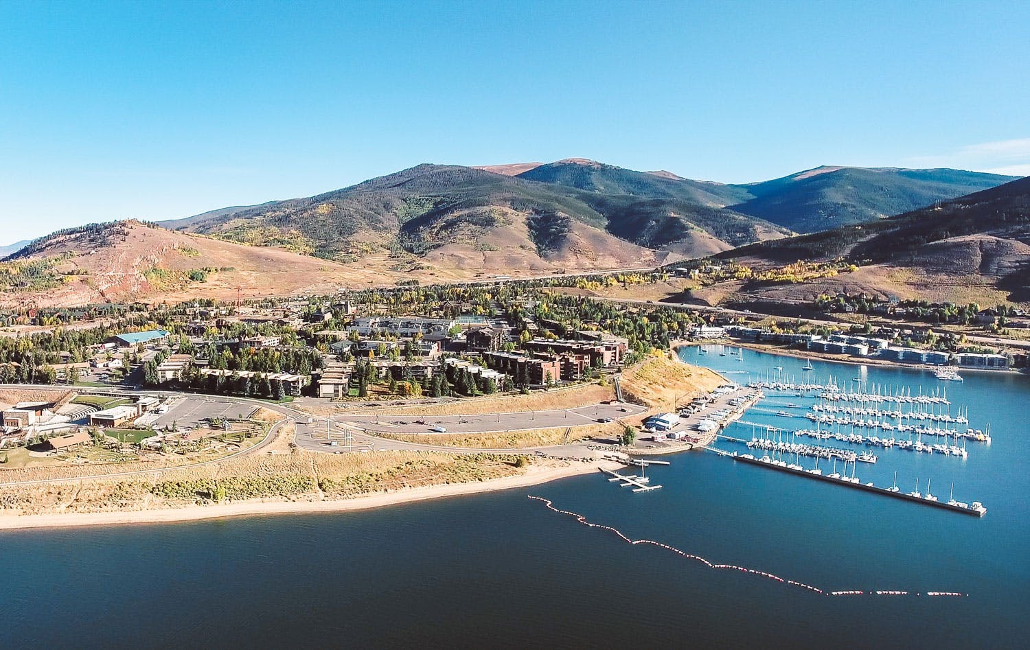 An aerial shot of a blue-sky day on Lake Dillon with the marina to the right and the town sits on the shores with yellow grass and evergreen trees.