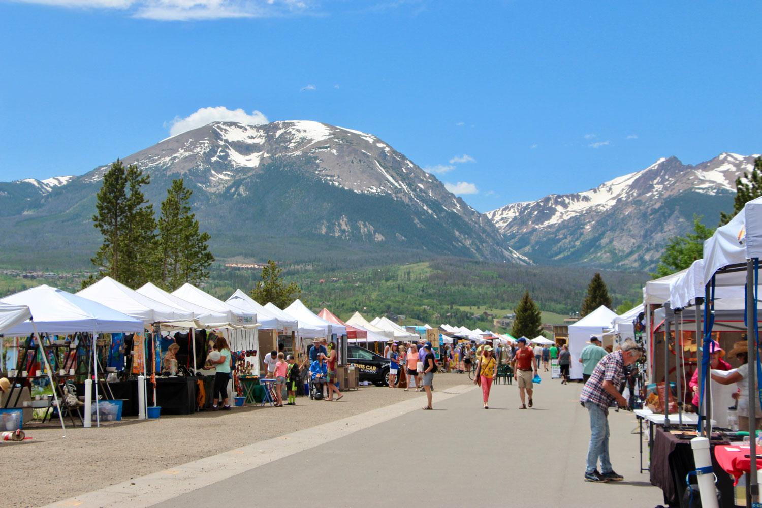 Beneath snow-capped mountains and a bright-blue sky the Dillon Farmers Market's white plastic tents sit on grey pavement. People walk in front of the booths that are on both sides.