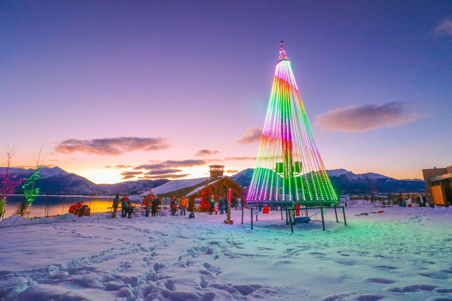 A rainbow lit up Christmas tree sits on a snowy field in Dillon Town Park at dusk. The sky is purple with pink and yellow hues as the sun sets behind the mountains.