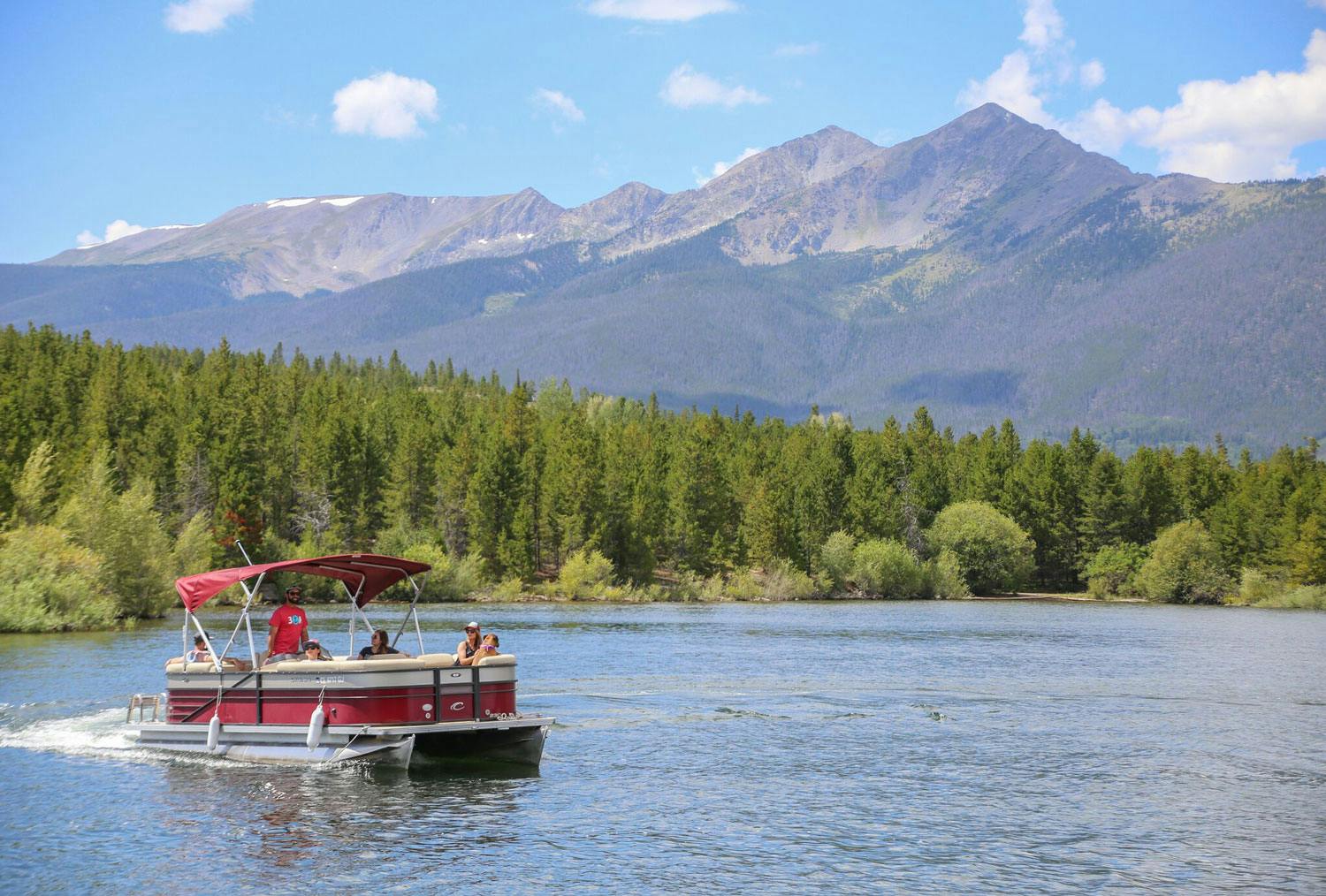 A red flat-bottomed boat makes its way across Lake Dillon with evergreen trees on the shore. In the background, mountain peaks rise up to the blue sky with white clouds.