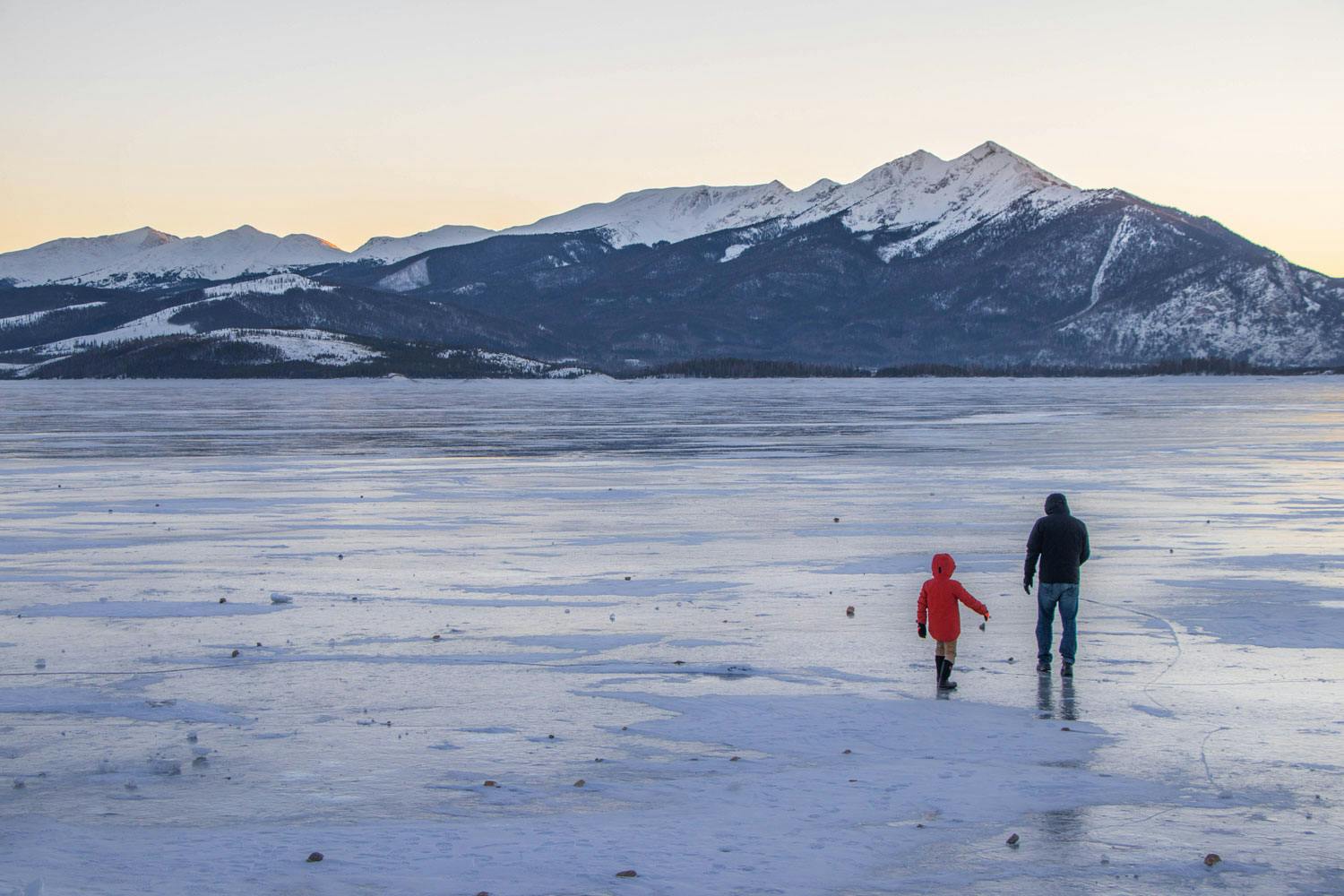 Two people, one in a red jacket, one in a black jacket walk across the frozen Lake Dillon. In the distance, snow-capped mountains sit in a light-yellow sky.