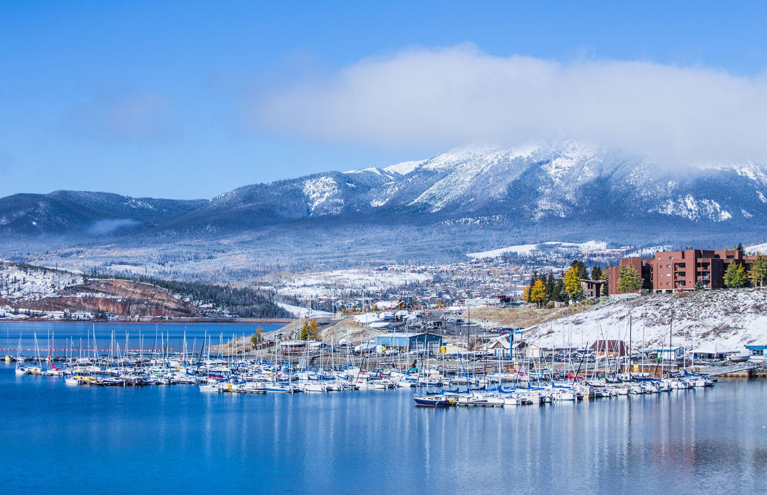 On the blue lake, the Dillon Marina sits with many boats on a winter's day. In the distance red-brick multi-story buildings sit on the shores and snow-covered mountains are covered slightly in clouds on a blue sky.