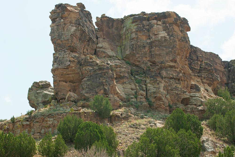 A photograph of a large, multi-tiered rock structure, painted different colors of brown, beige and green from time and weather. Below the rock structure, the Windsplitter Rock at the Wild Animal Refuge in Springfield, Colorado, are several green bushes.