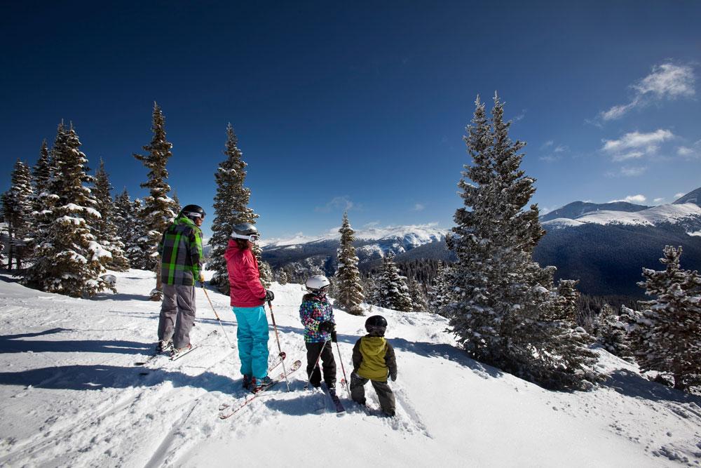 Two adults and two kids on skis stop to soak up the view of snowy peaks under a bight-blue sunny sky.