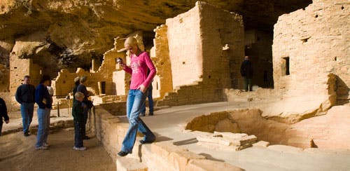 A person in a pink top with blue jeans walks away from a sand-colored cliff dwelling.