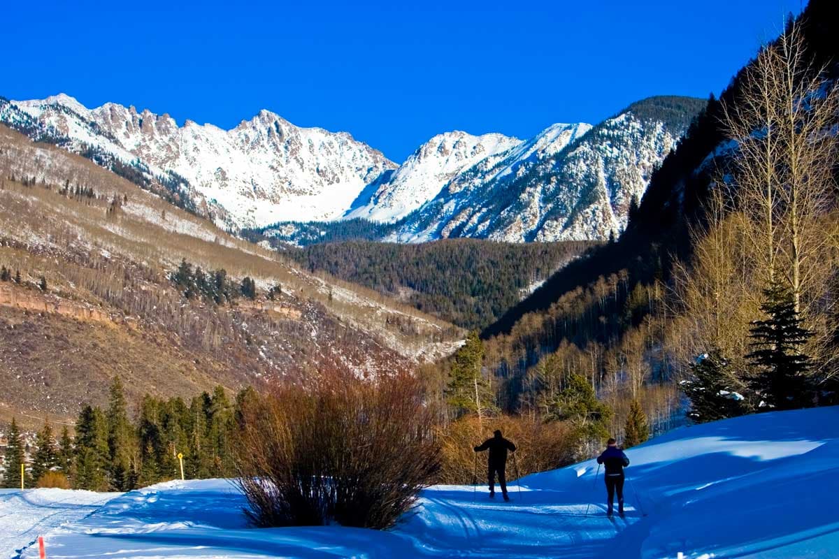 Two cross-country skiers traverse a shaded area of hilly, groomed trail in the shadow of snow-capped mountains near Vail, Colorado