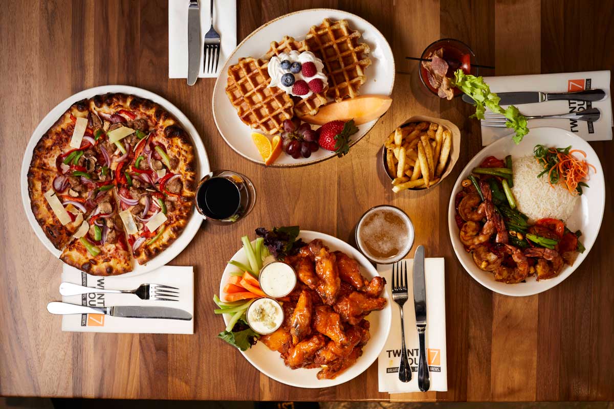 Table with plates of waffles with whipped cream, spicy shrimp and rice, chicken wings and veggies, and a pizza.