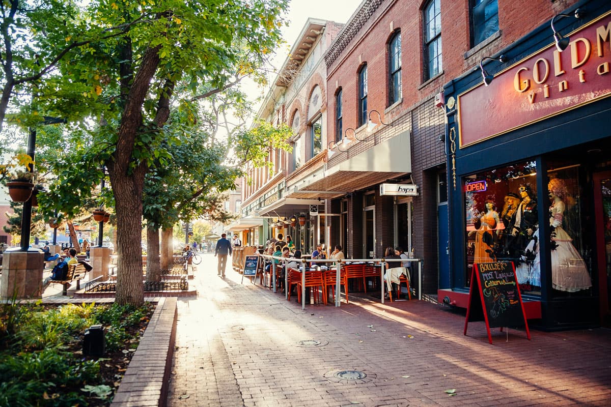 A row of trees behind low brick walls create shade from the summer sun for pedestrians walking along the boutiques of Boulder's Pearl Street.
