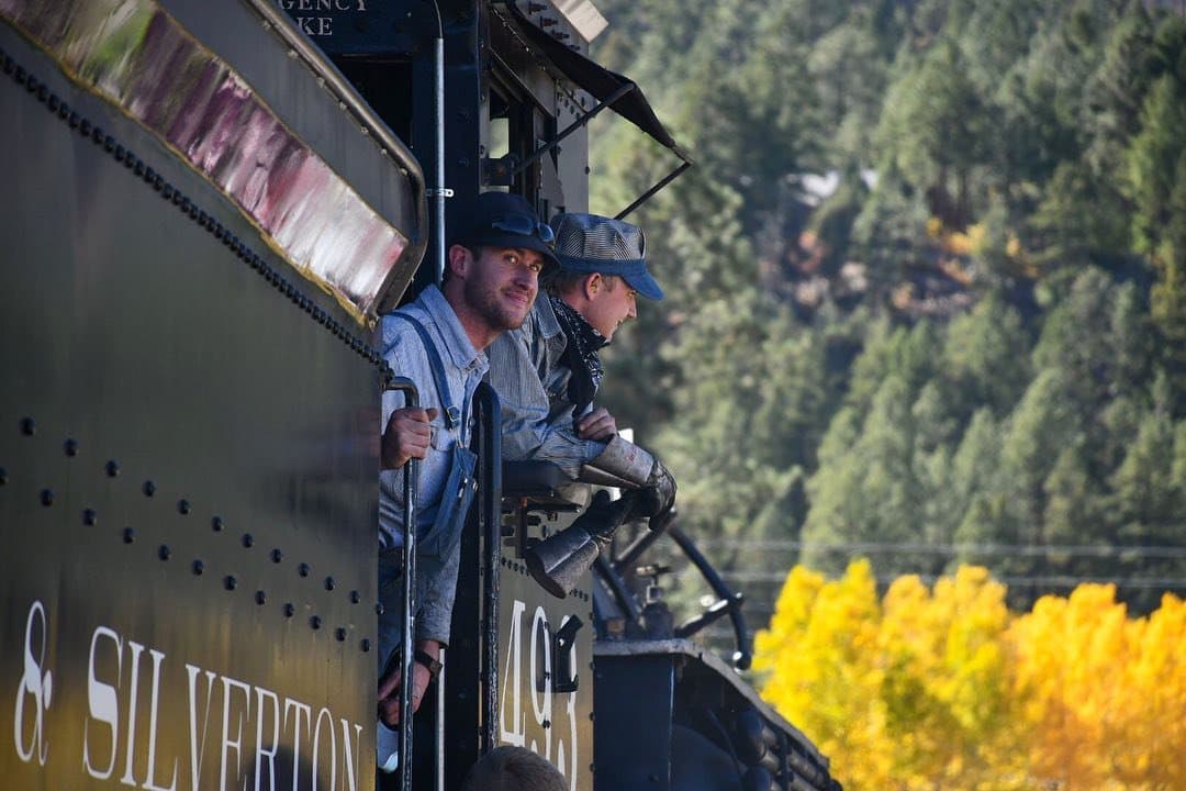 Two train engineers in historical 1800s dress lean out slightly from the engine room of the train in front of a golden stand of aspens. One engineer smiles at the camera.