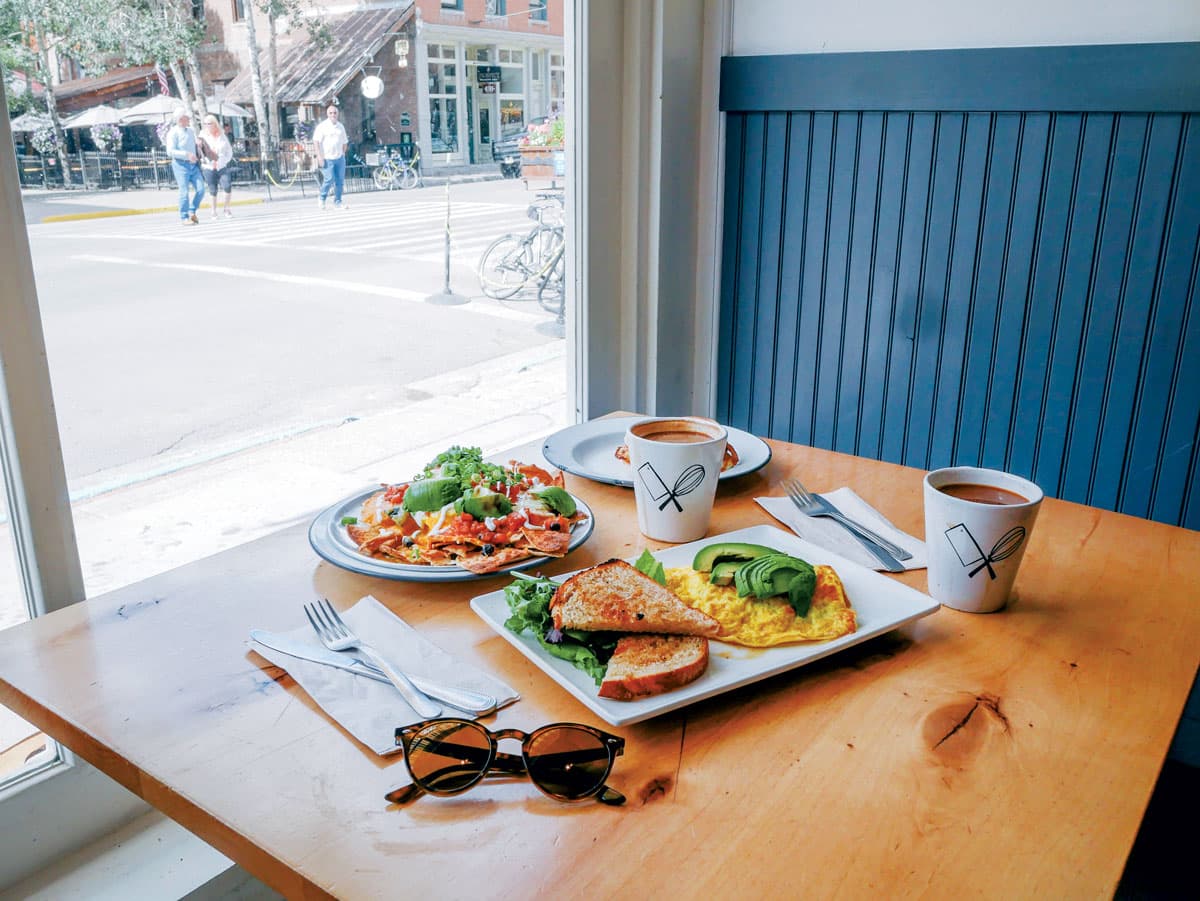 Two mugs of coffee and savory breakfast foods sit on a table near a large window inside The Butcher & The Baker in Telluride, Colorado.