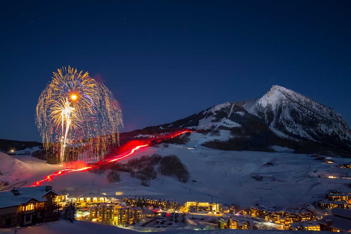Fireworks over Crested Butte Mountain Resort. We see hotels and restaurants lit up, a timelapse of thorchlight moving down the mountain carried by skiers.