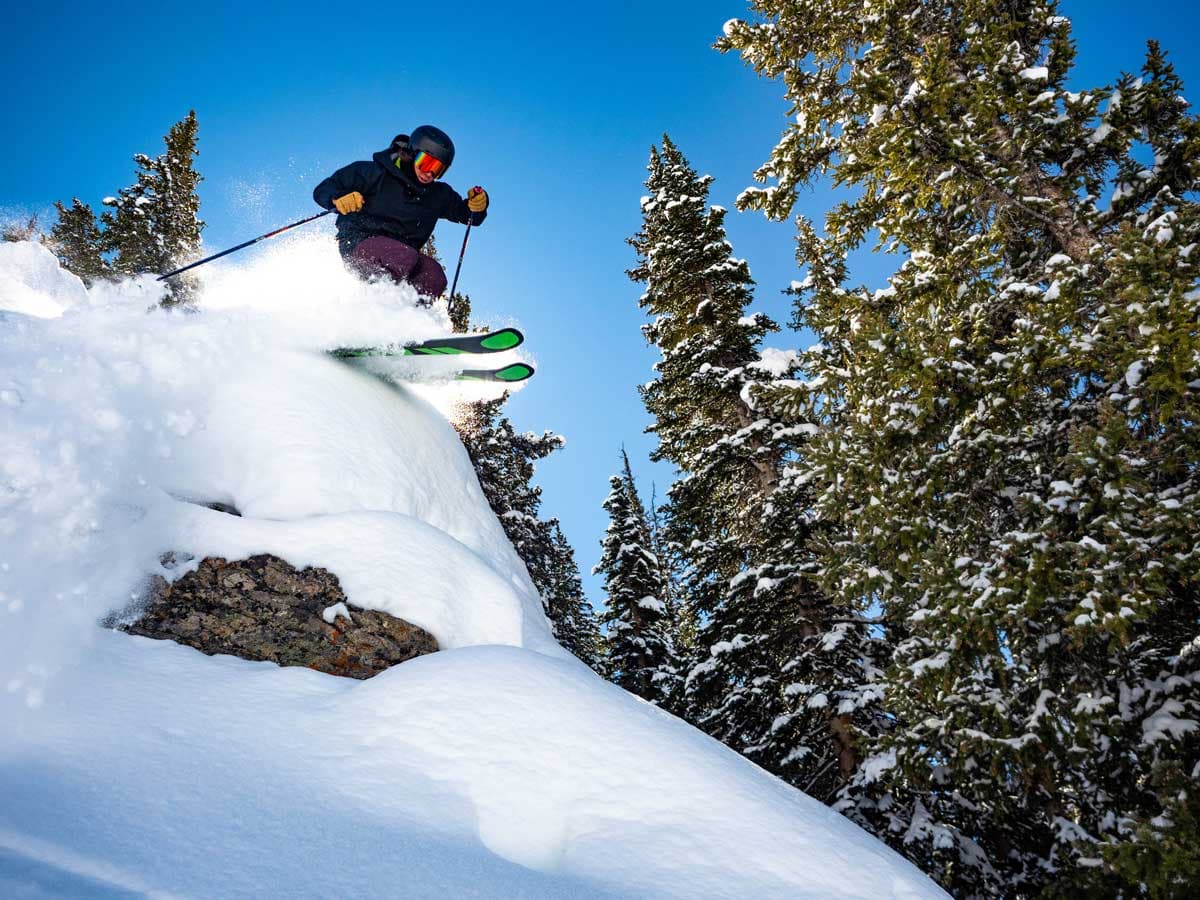 A skier flies through the air over a rock covered in feet of fluffy snow, with snowcovered trees around them