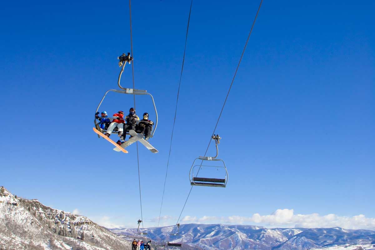 Snowboarders and skiers ride a chairlift at Snowmass Mountain with a vast bluebird blue sky behind them