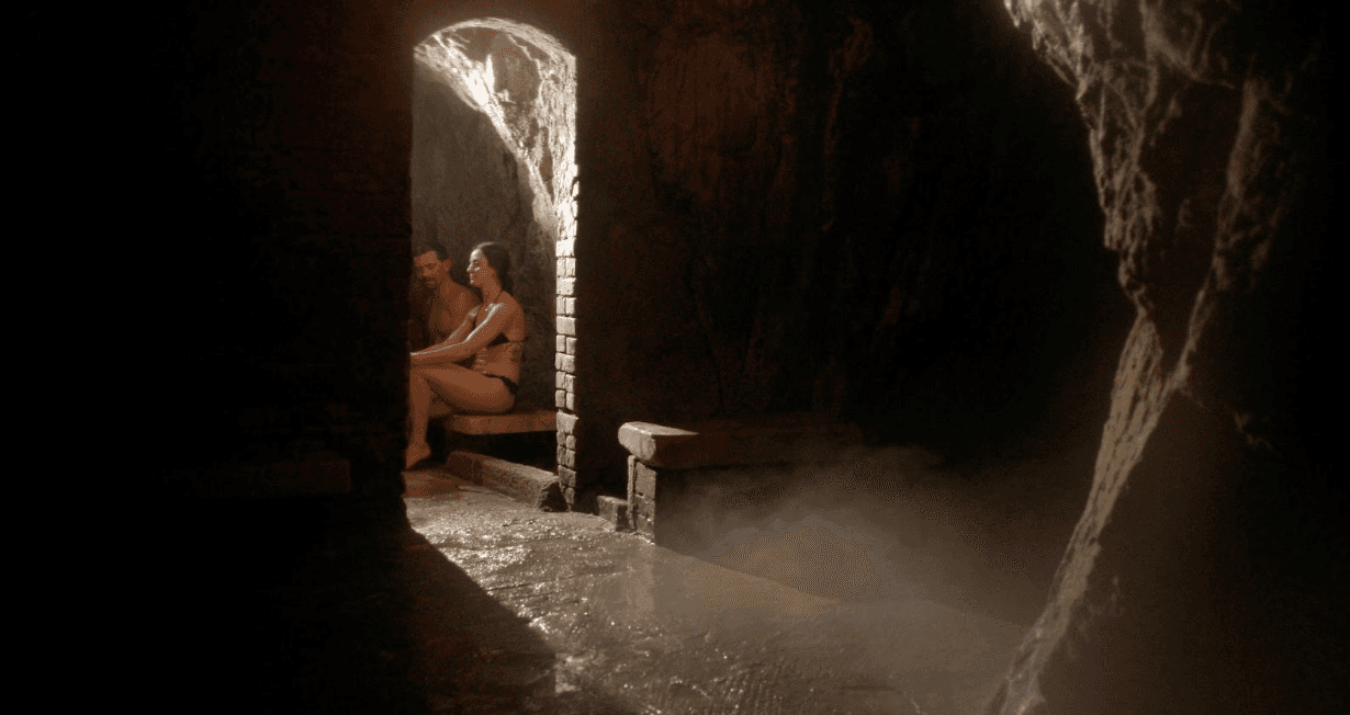 A couple wearing swimsuits sits on a wooden bench in a dimly lit cave and breathe in the steamy vapors at a spa in Colorado.