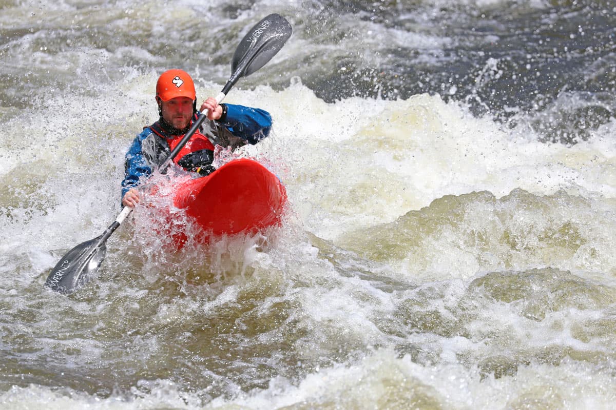 A solo kayaker with a red helmet on sitting in a red kayak is cruising down a whitewater river. They have their paddle in hand and are paddling through the water as the kayak is moved by the active water.