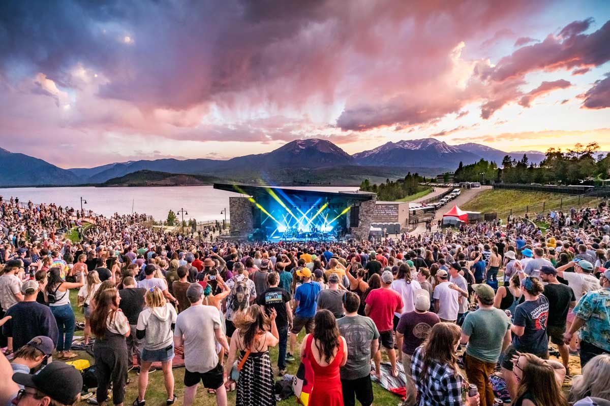 A crowd listens to a band perform from a blue-lit stage with Dillon Reservoir, mountain peaks and pink clouds as the backdrop