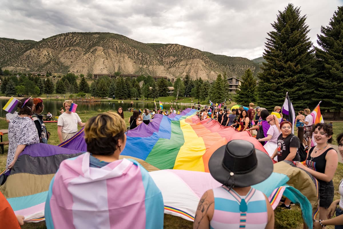 Mountain Pride celebrators stand next to a small lake, dressed in various pride flags. In the middle is a rainbow pride flag held by the people surrounding it near Avon.