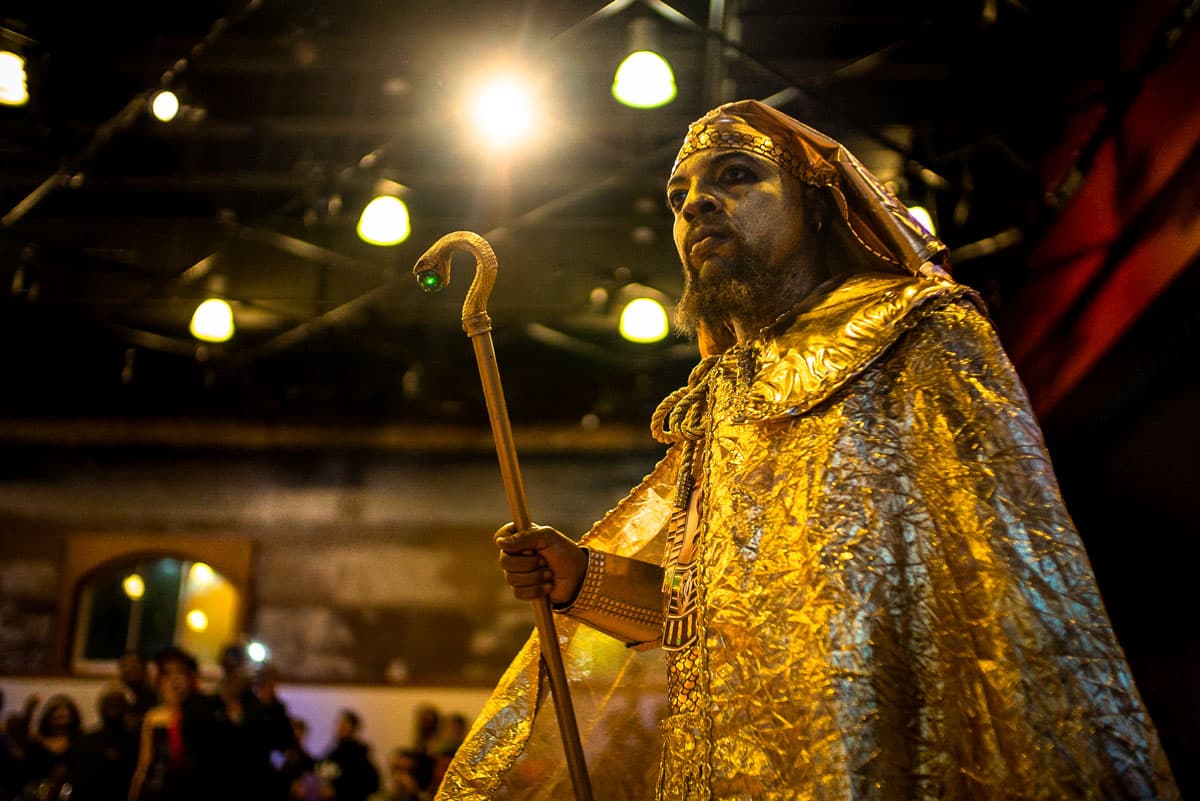 A person stands under stage lights dressed like a golden Pharaoh, complete with golden face paint and a golden cape. They hold a staff in their hand.