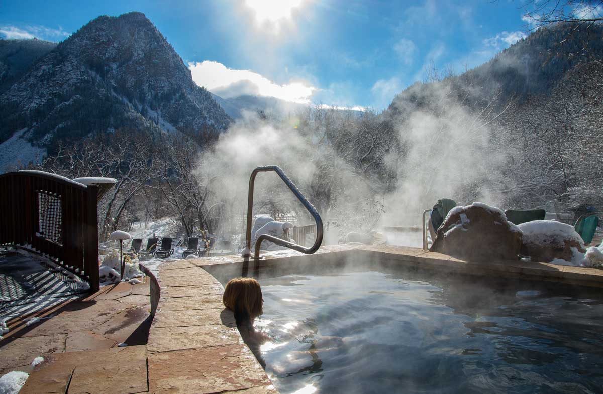 Steam rises from a heated hot spring pool at Avalanche Ranch in Colorado. A person soaks in the water and rests their head against the sandstone ledge.