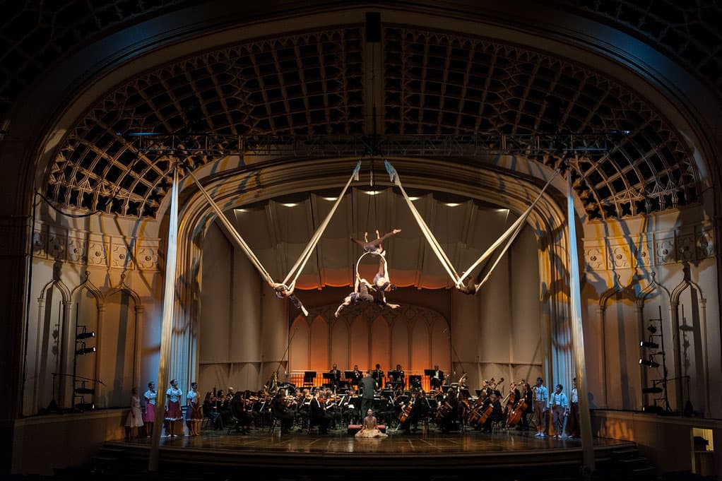 A stage with philharmonic performers playing music. Above them, aerial performers defy gravity