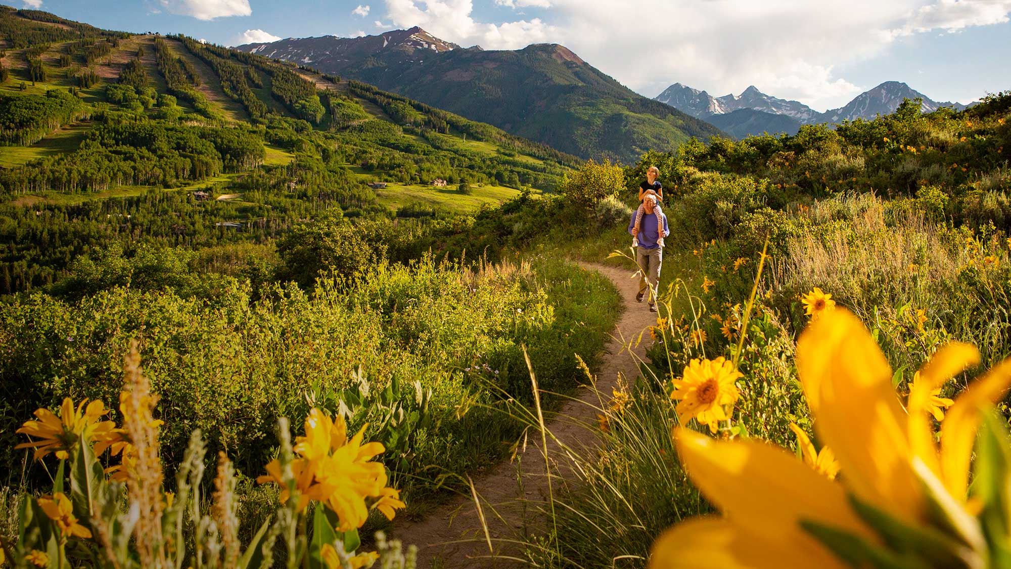 A man carrying a child on his shoulders walks on a path surrounded by wildflowers and mountain peeks