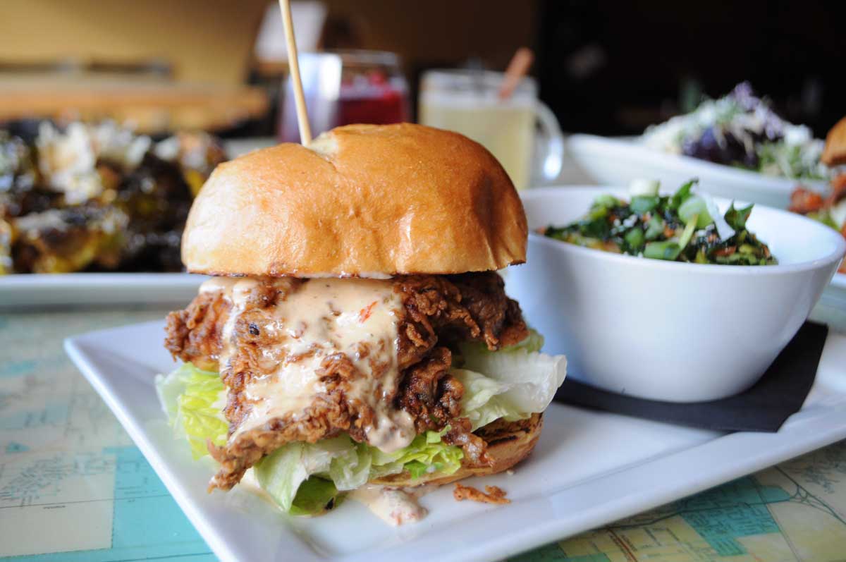 A fried-chicken sandwich with all the toppings on a white plate next to a salad-filled salad bowl