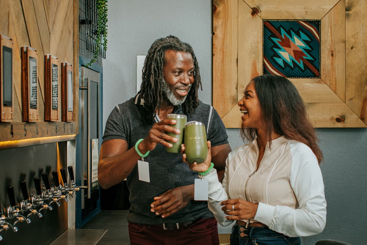 Two smiling people cheers their dark-green tumblers at the Oakwell Beer Spa taproom. In the background to the left of the photo are individual taps which feature beers from local Colorado breweries.