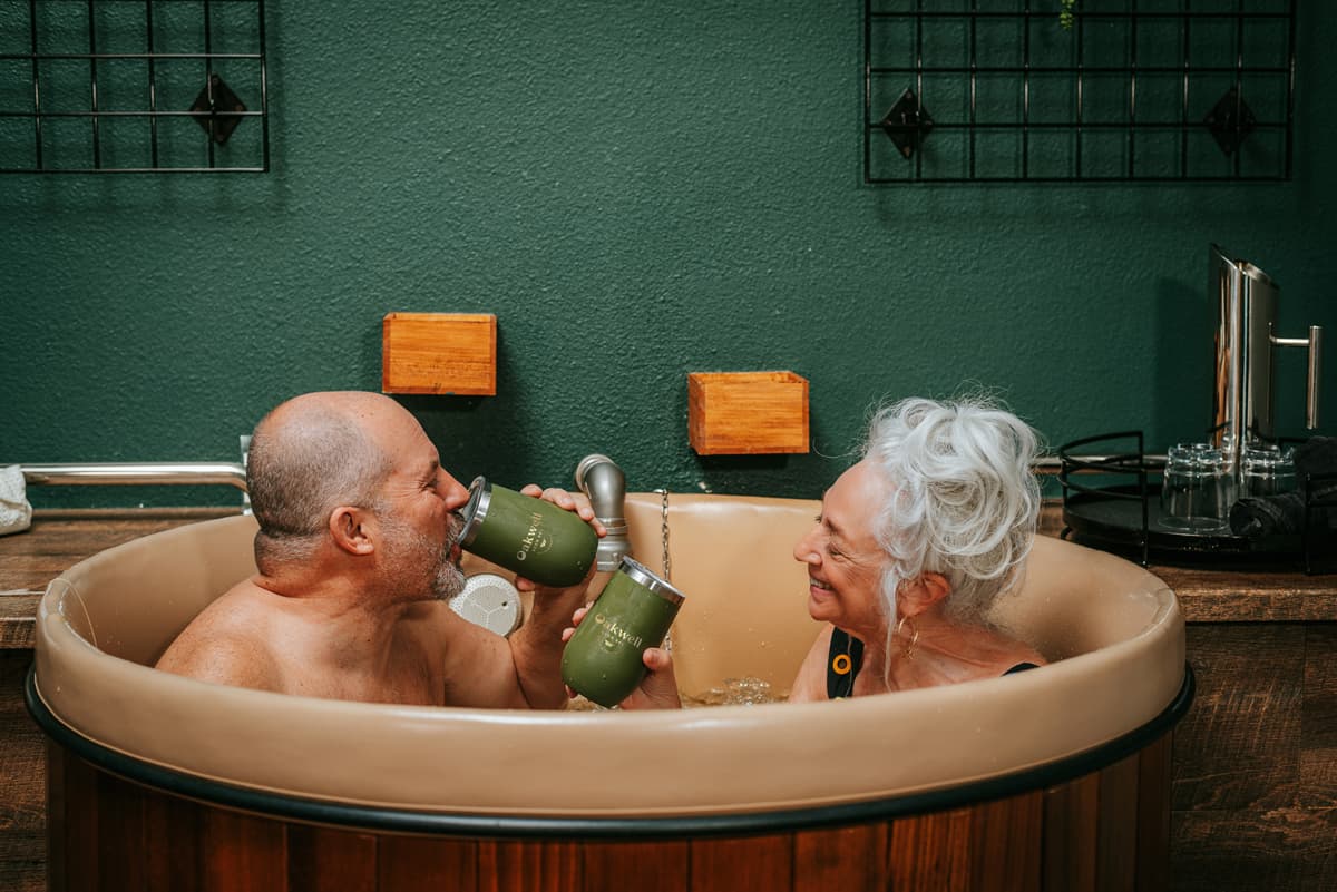 Two older people smile and drink from their Oakwell Beer Spa tumblers in a Hydrotherapy Beer Bath. The background of the room is painted a soothing dark green.