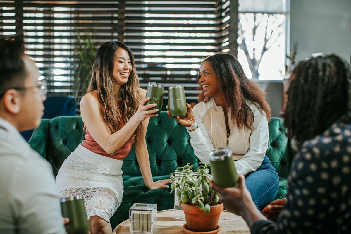Four smiling and laughing friends drink beer at Oakwell Beer Spa in the taproom with their beer tumblers. The sun shines through windows behind them.