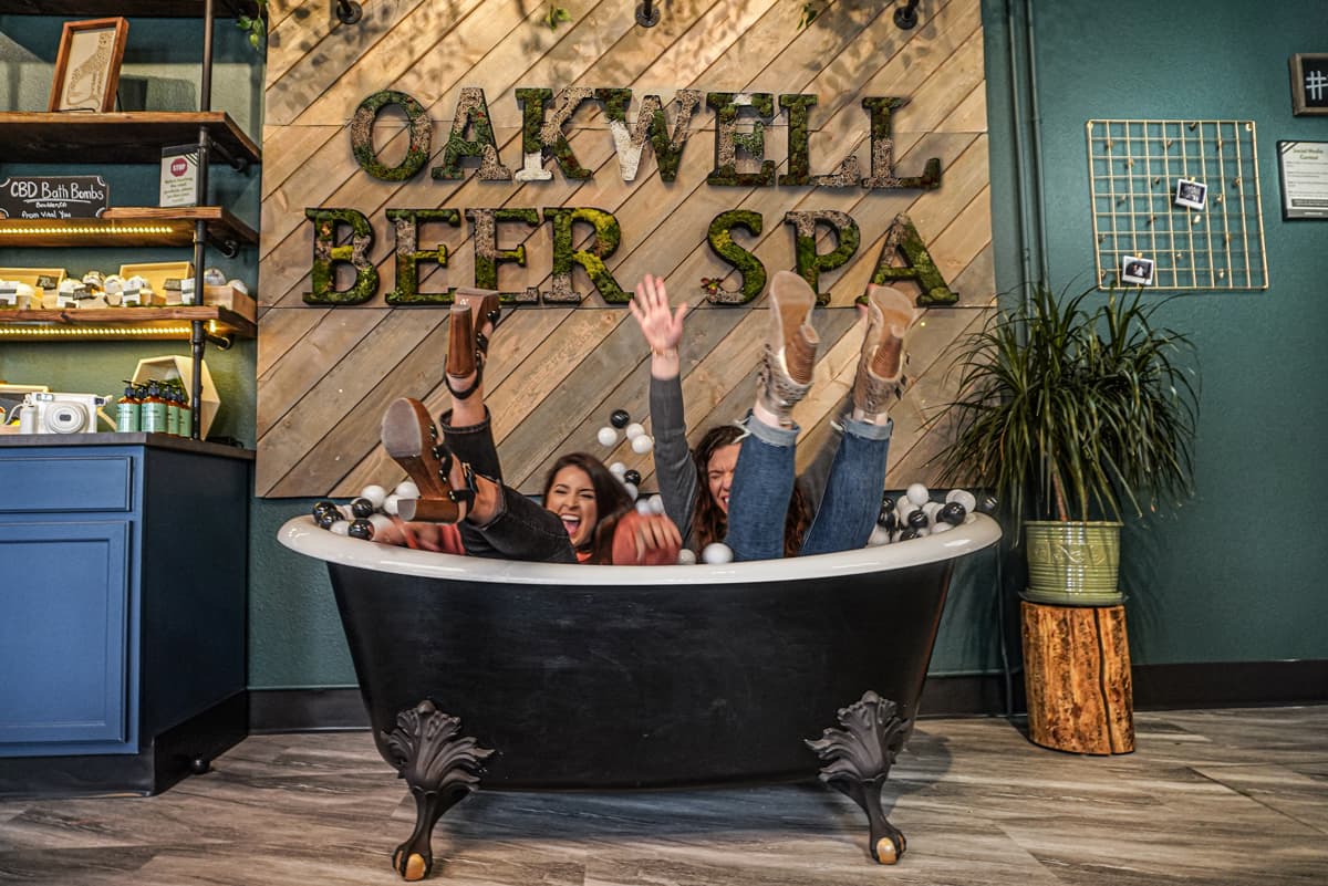 Two laughing friends fall backwards into the vintage-looking clawfoot tub under a large sign that says "OAKWELL BEER SPA" in all caps. There are pastel-colored balloons floating around them and in the tub.