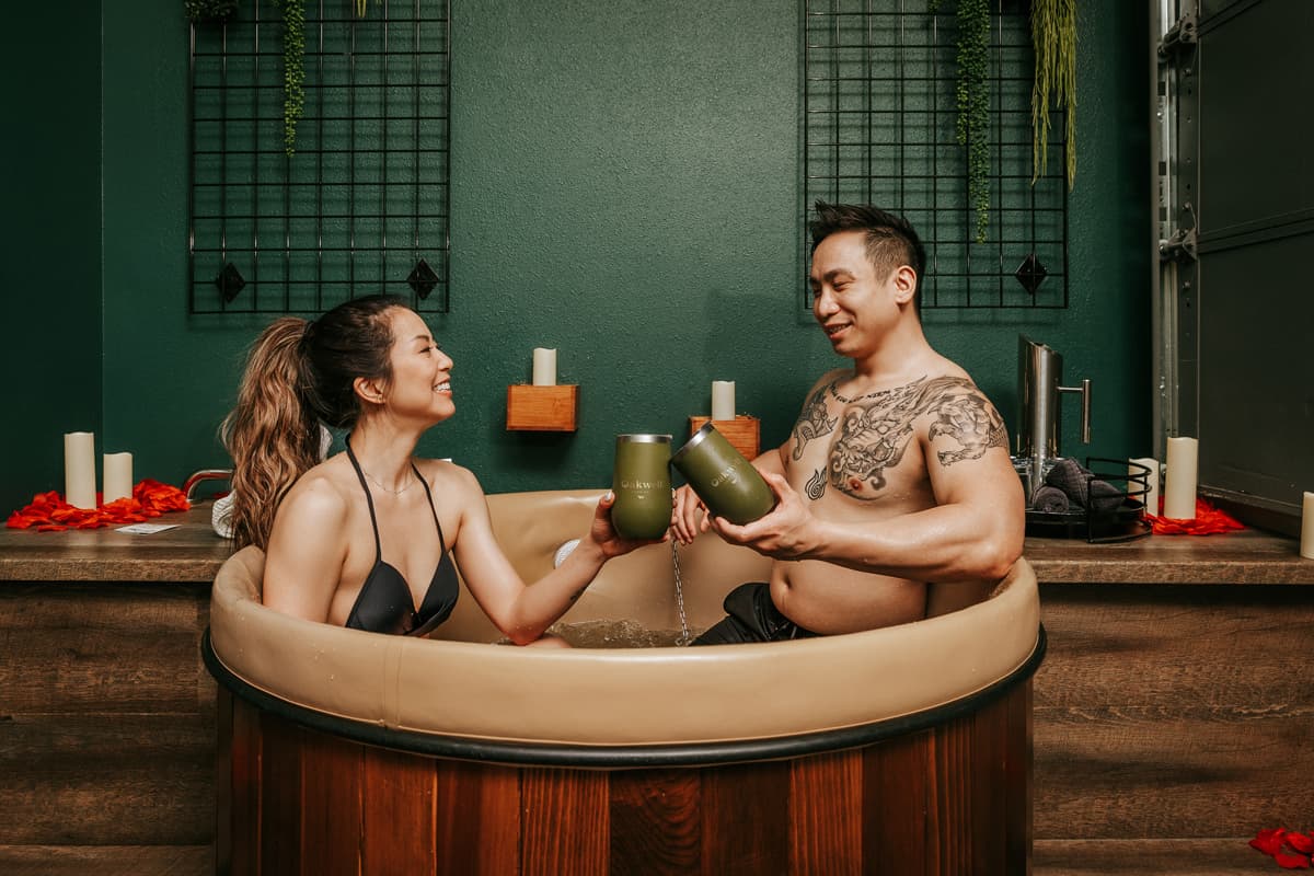 A couple standing in the beer bath at Oakwell Beer Spa smile and laugh as they face each other and cheers their tumbler full of local Colorado beer. In the background are tall candles and rose pedals, as part of the spa's Valentine's Day special.