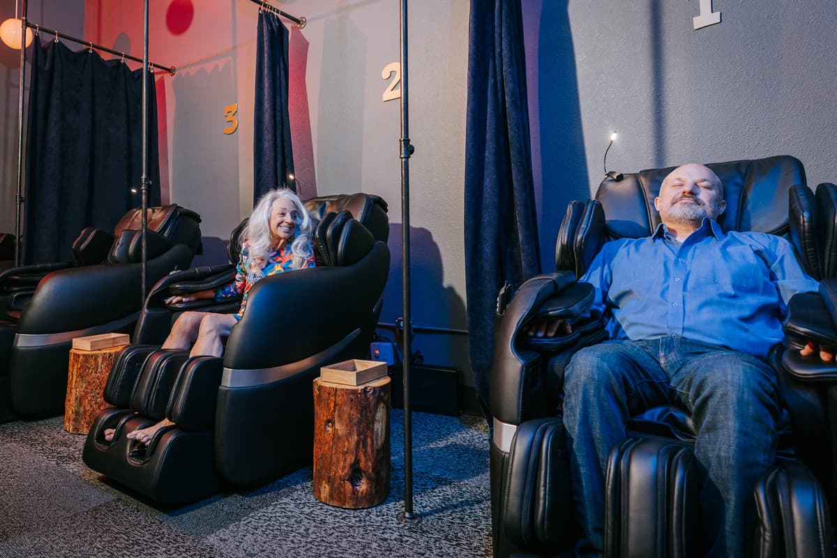 Two people smile while sitting in the zero-gravity massage experience at Oakwell Beer Spa.The room is darkly and serenely lit, and the people in the chairs look relaxed. There are black curtains to separate each massage unit for ultimate privacy.