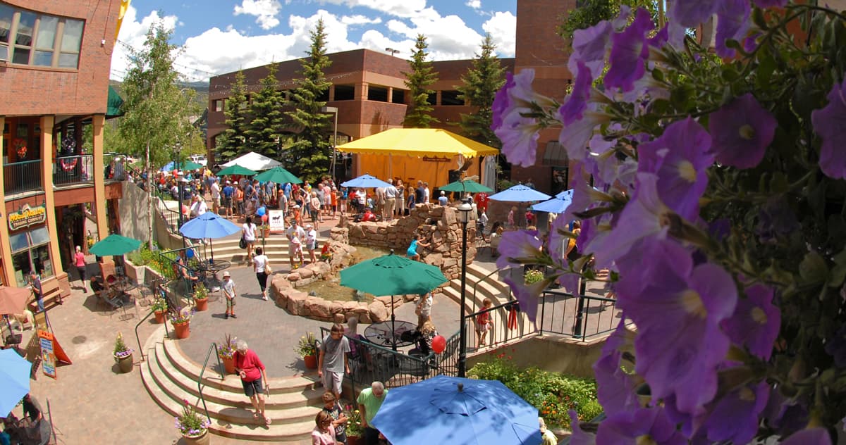 A busy courtyard with sidewalks and circular walking paths is full of people enjoying the summer time in Winter Park, Colorado. There are trees and flowers all around.