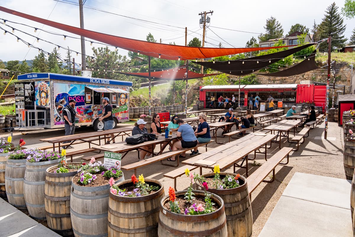 People sit at picnic tables under canopies on a patio lined with food trucks and wooden barrels filled with colorful flowers at The Barrel in Estes Park, Colorado.