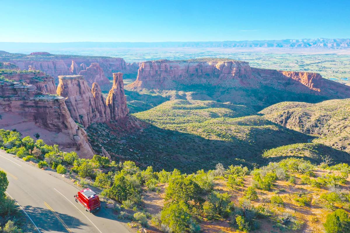 In the bottom left of the photo is a large red van driving along Rim Rock Drive in Colorado National Monument in Grand Junction, Colorado. The scene on the right of the van is full of vast desert landscapes, with several plateau-looking towering rock formations. The sky is clear and blue above the whole scene.