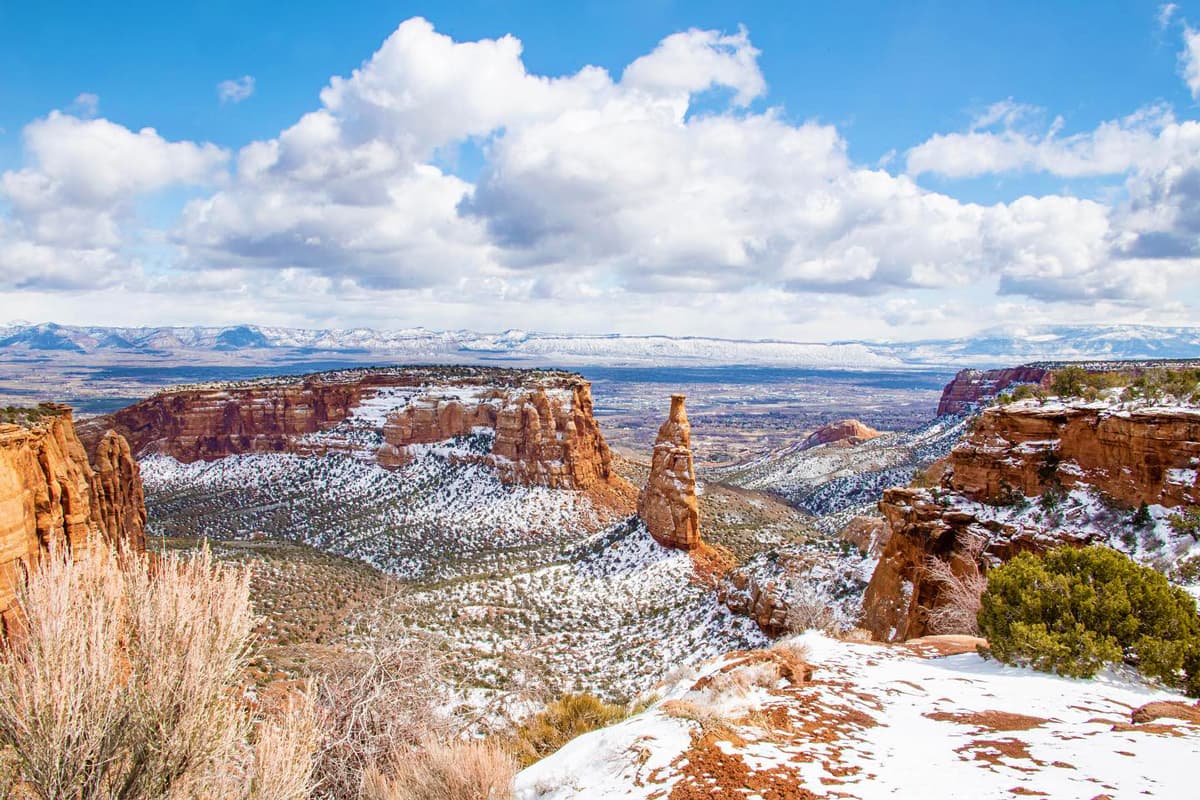 An aerial view of Grand Junction, Colorado in the winter. There is a light dusting of snow on jutting stone formations across the landscape and in the background are more formations and vistas of mountains. The cloud are puffy and gray above the scene.