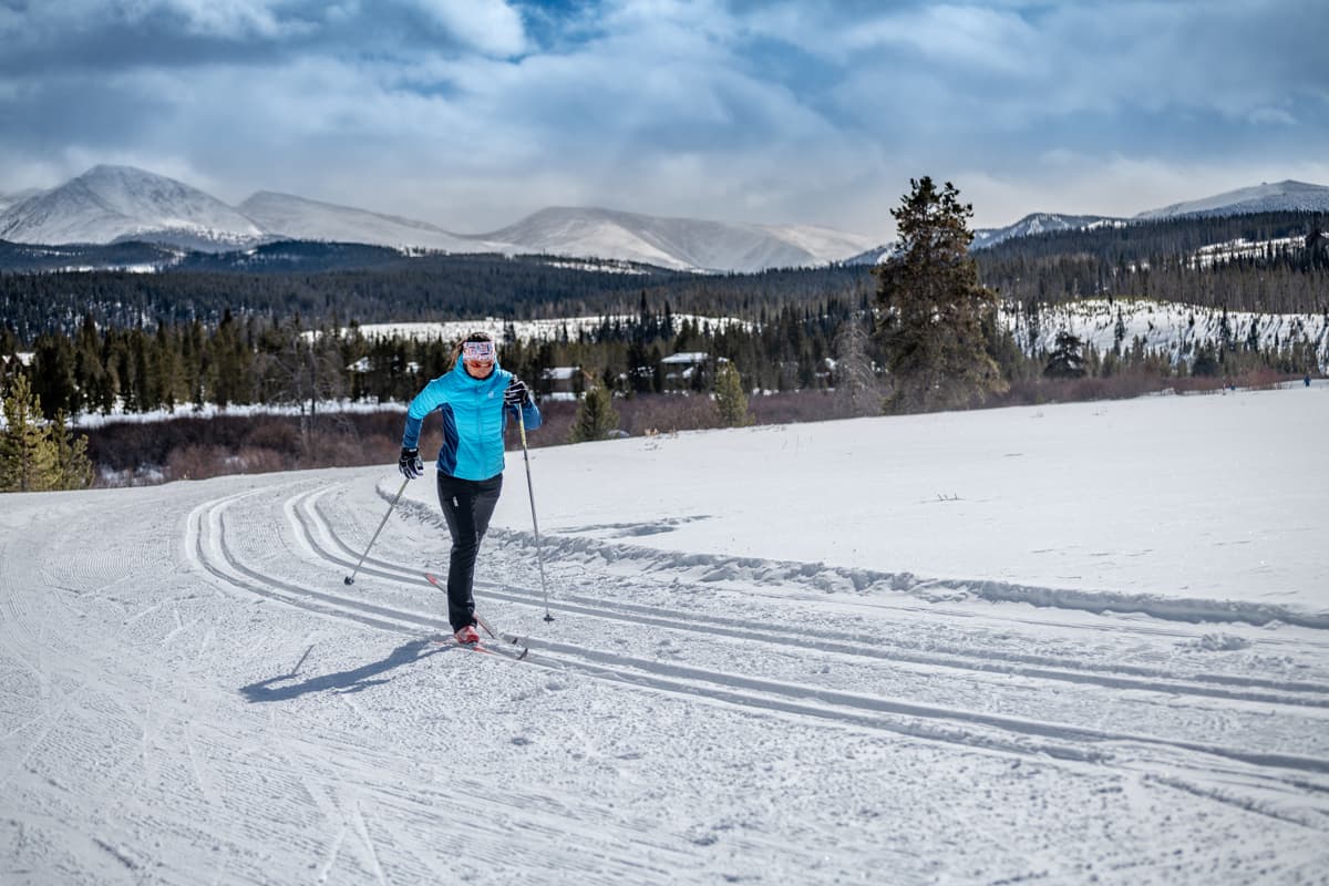 A lone skiier glides along groomed ski tracks at a Nordic center in or around Winter Park. They are dressed in warm clothing as they scoot along the tracks. All around them are hills laden with snow and tall pine trees. In the distance, clouds cover the tops of snowcapped mountains.
