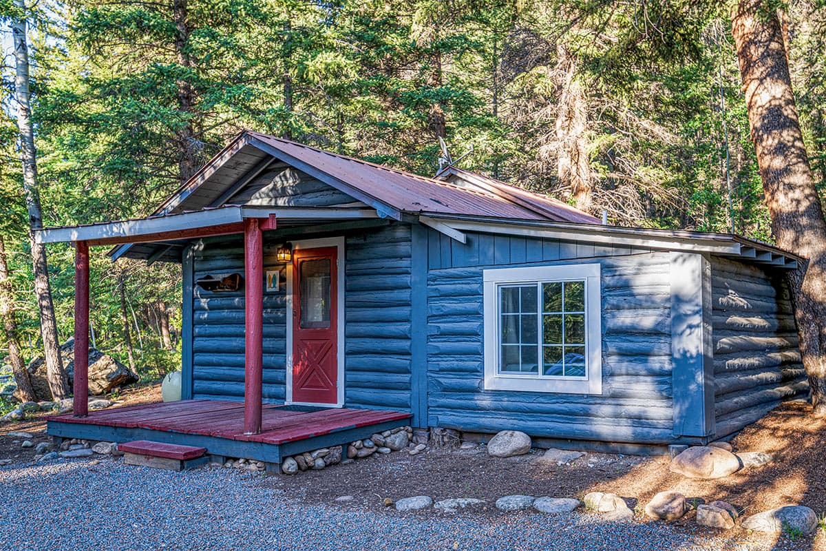 A blue cabin lies on the Harmels on the Taylor property. It has a small porch. Pine trees stand tall behind it.