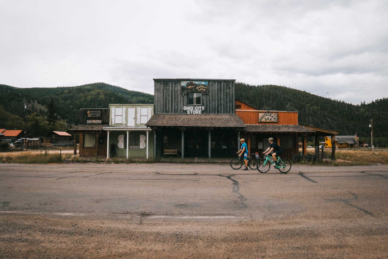 Two people gravel bike on teh rough road in front of the ghost town facades of a store and restaurant in Ohio City near Crested Butte