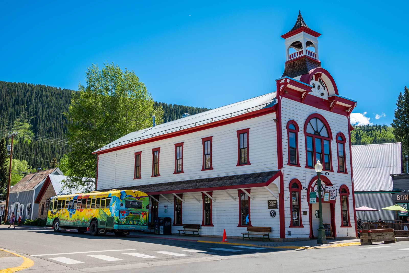 A yellow bus with a leafy mural stops beside a historical white-and-red building