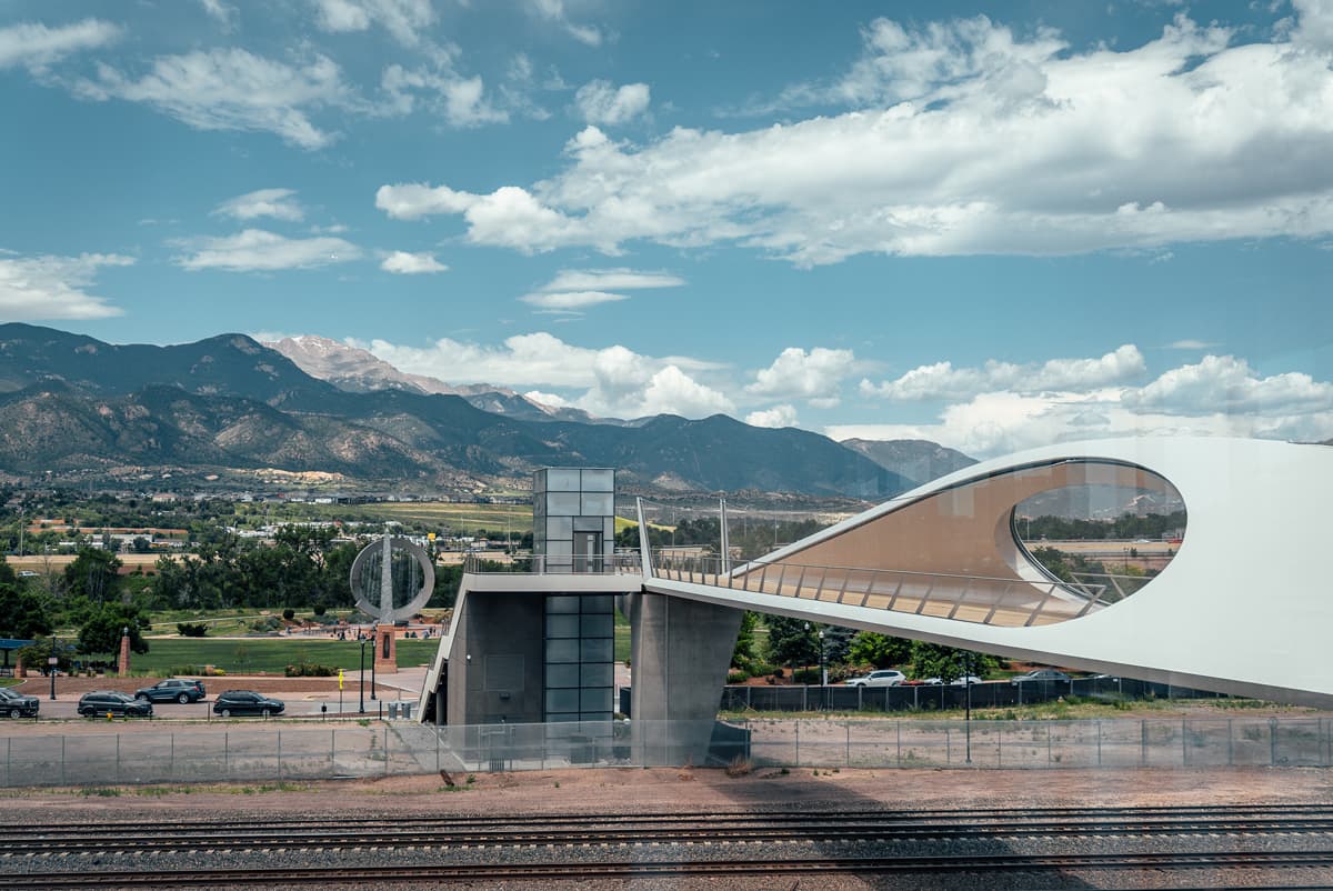 A futuristic bridge over train tracks leading to a park with a large silver sculpture with water feature and Pikes Peak in the background at  US Olympic & Paralympic Museum  in Colorado Springs